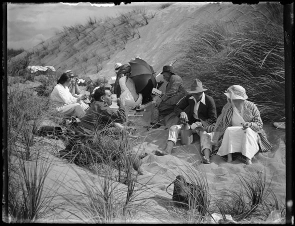 Black and white photograph of a group of people sitting in sand dunes, eating and drinking.
