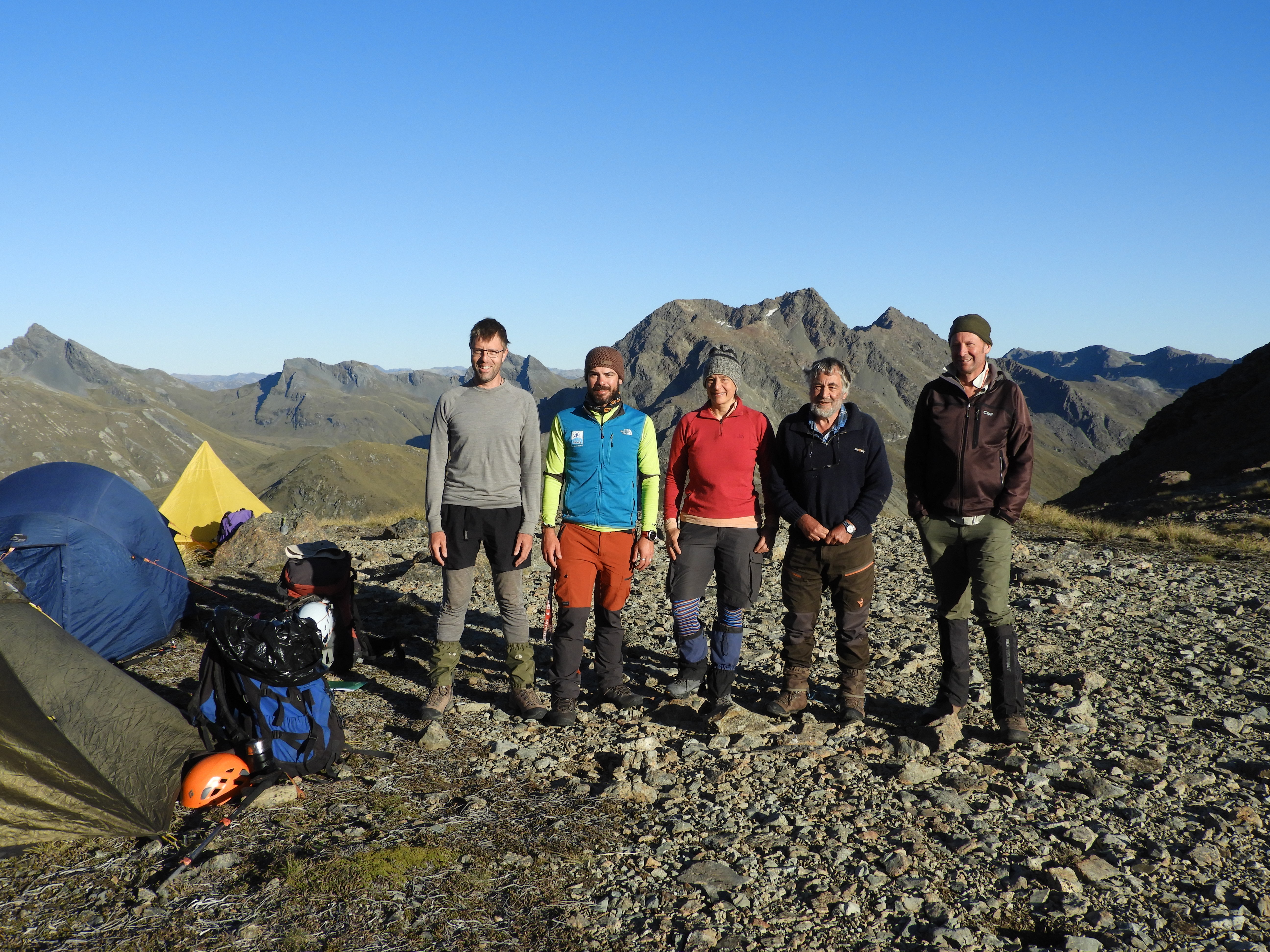 Five people in hiking gear standing next to tents with mountain ranges and blue sky behind them