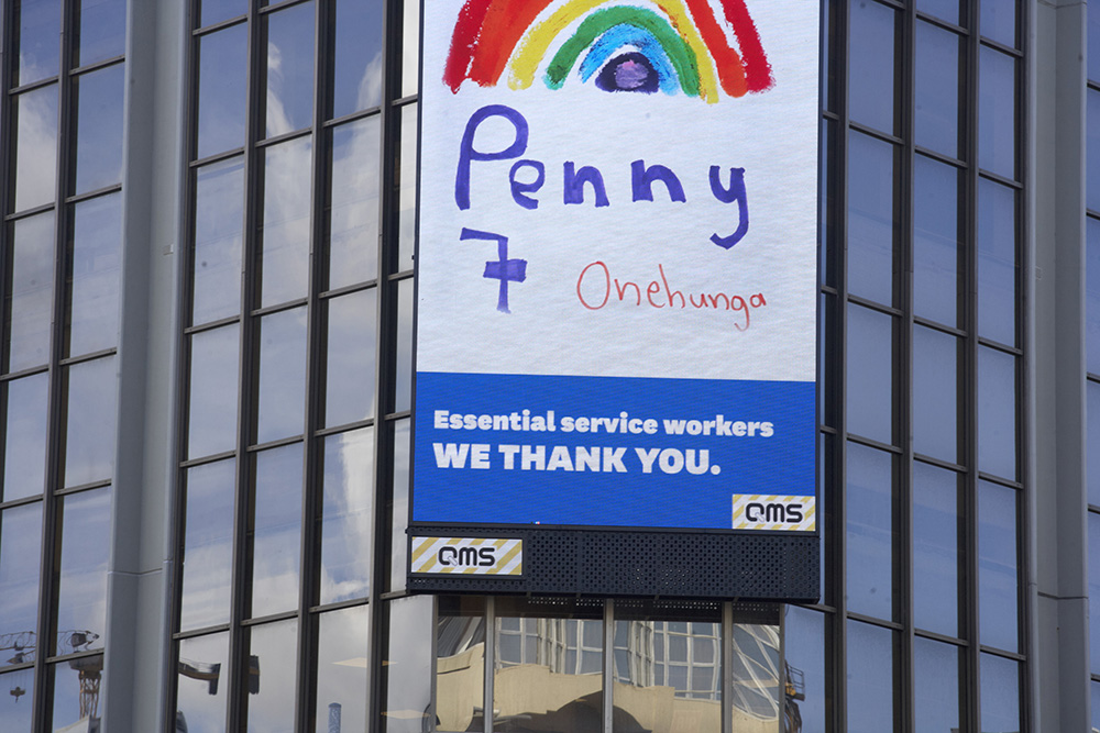 Close up on a digital sign on the sie of a building. The sign is displaying a poster of a hand-drawn rainbow, with the words ‘Essential service workers we thank you’
