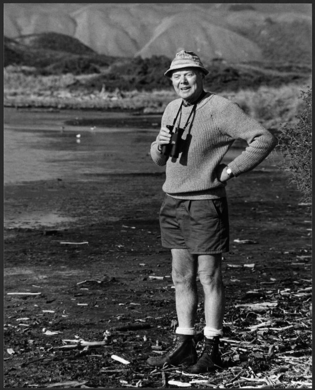A black and white photo of a man in shorts and a hat standing next to a lagoon holding binoculars