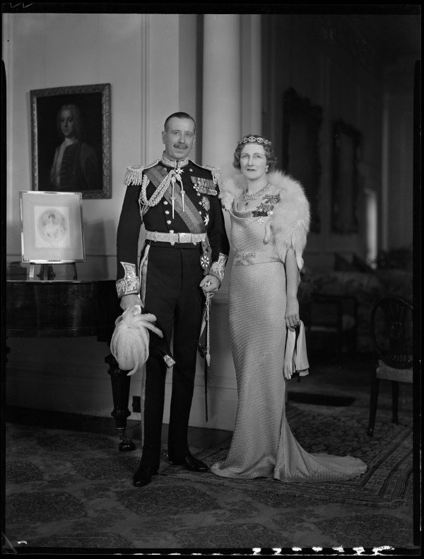 A black and white photo of a couple standing in a large room. The man is in Army dress suit and the woman is dressed in fine clothes and wearing a tiara.