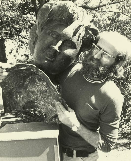 A photograph of a bearded man lifting a sculpture of a head off a plinth