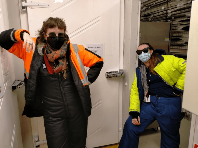 Two women in sunglasses, facemasks and cold-weather gear stand casually in an inside doorwayy
