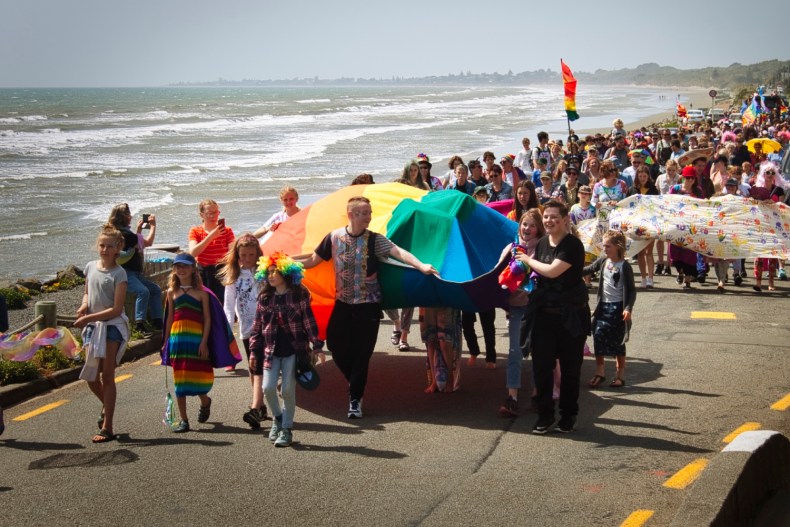 A colourful parade of adults and children walking along a coastal road with the sea in the background. The people in front are holding the edges of a large rainbow flag.