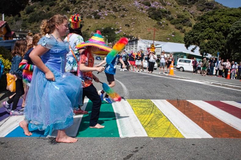 Children in colourful fancy dress costumes are crossing a pedestrian crossing with filled in with rainbow colours. There is a hill in the background and people watching on.