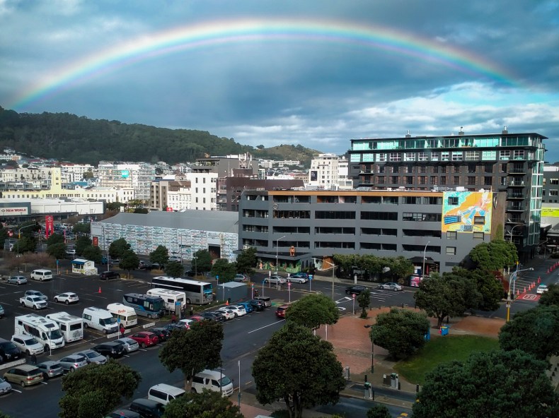 A photo of a full rainbow stretching over a section of Wellington. There is a carpark and trees in the foreground and buildings and a hill in the background.
