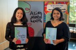 Grace and Mehwish stand in front of a sign advertising the Asian Mental Health project, holding copies of the zine