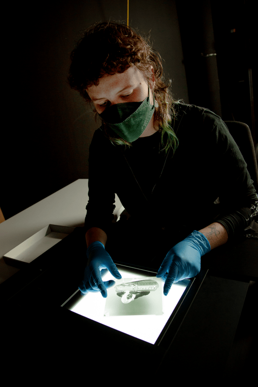 A dark photo of a person wearing a facemask and blue gloves holding a photographic negative on a lightbox. 