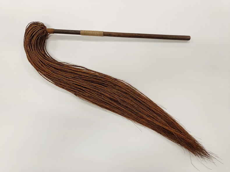 A short wooden stick with a long tassel of woven threads coming off one end of it.