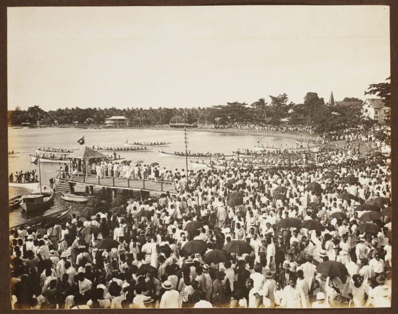 A sepia photograph of a large crowd of people standing at the water's edge next to a pier. There are several long canoes in the water with many rowers in each.
