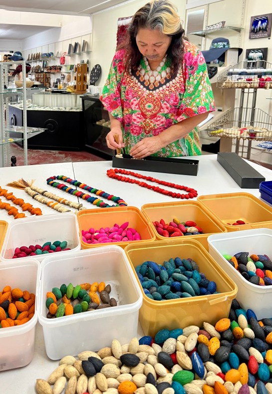 A woman in a brightly coloured top puts an equally brightly coloured karaka berry necklace into a box. In the foreground are ice-cream containers with all the different coloured berries sorted into each one.