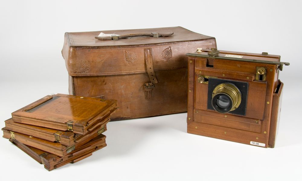 A wooden camera, leather camera box, and wooden glass plate holders arranged on a white surface.