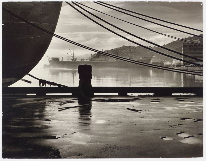 An artistic black and white photograph of a the end of a ship (unclear which end) tied to a wharf with ropes on a rainy day. There is another ship in the mid-distance and the hills of the harbour in the far distance.