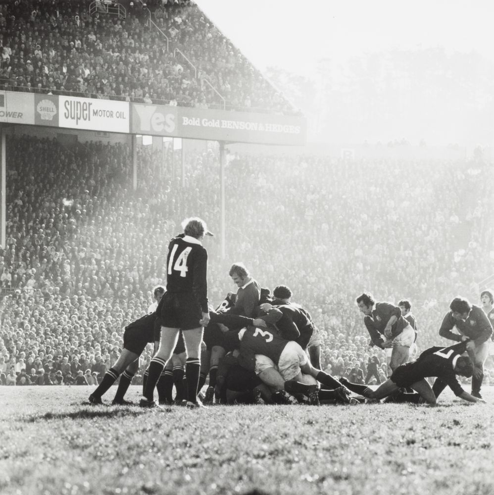 A football field with a full grandstand obscured by sun haze has two rugby teams in a scrum. One man is standing with his arm up sheilding his eyes from the sun.