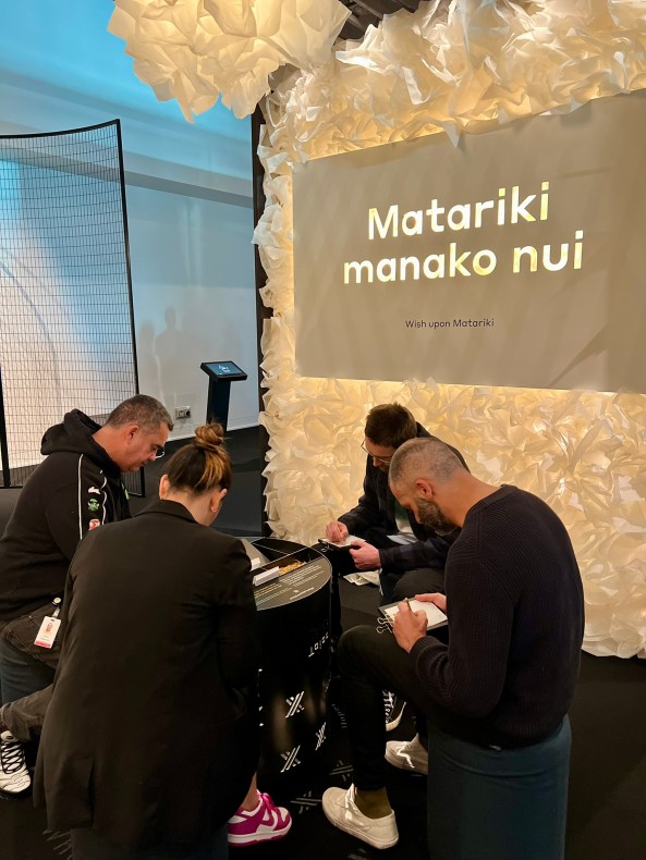 Four people in black sitting around a small table writing on A6 sized white paper. Behind them is glowing white wall with the words 'Matariki manako nui/ Wish upon Matariki'