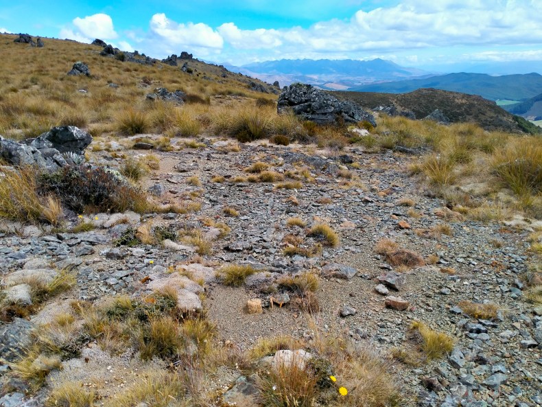 Rocky ground on top of a mountain. There are other mountains and cloudy blue skies in the distance.