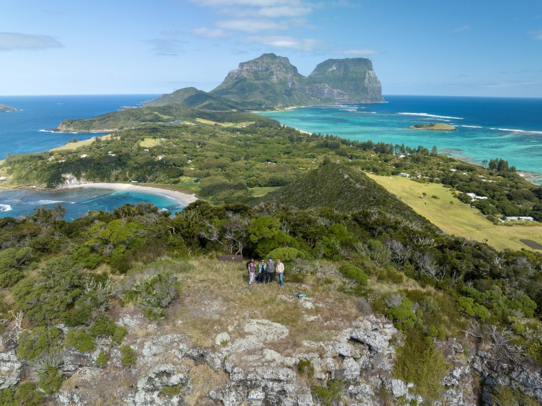 Photo from the air of four people standing together on a rocky outcrop. Out behind them is an island surrounded by the sea. There are rocky cliffs at the far end of the island. 
